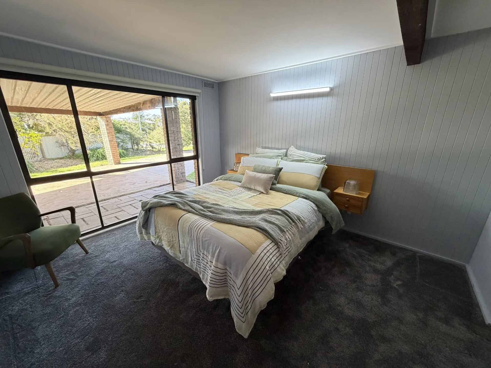 Master bedroom with a made-up queen bed, bedside tables, a green armchair and a large window looking out to a covered paved area and garden