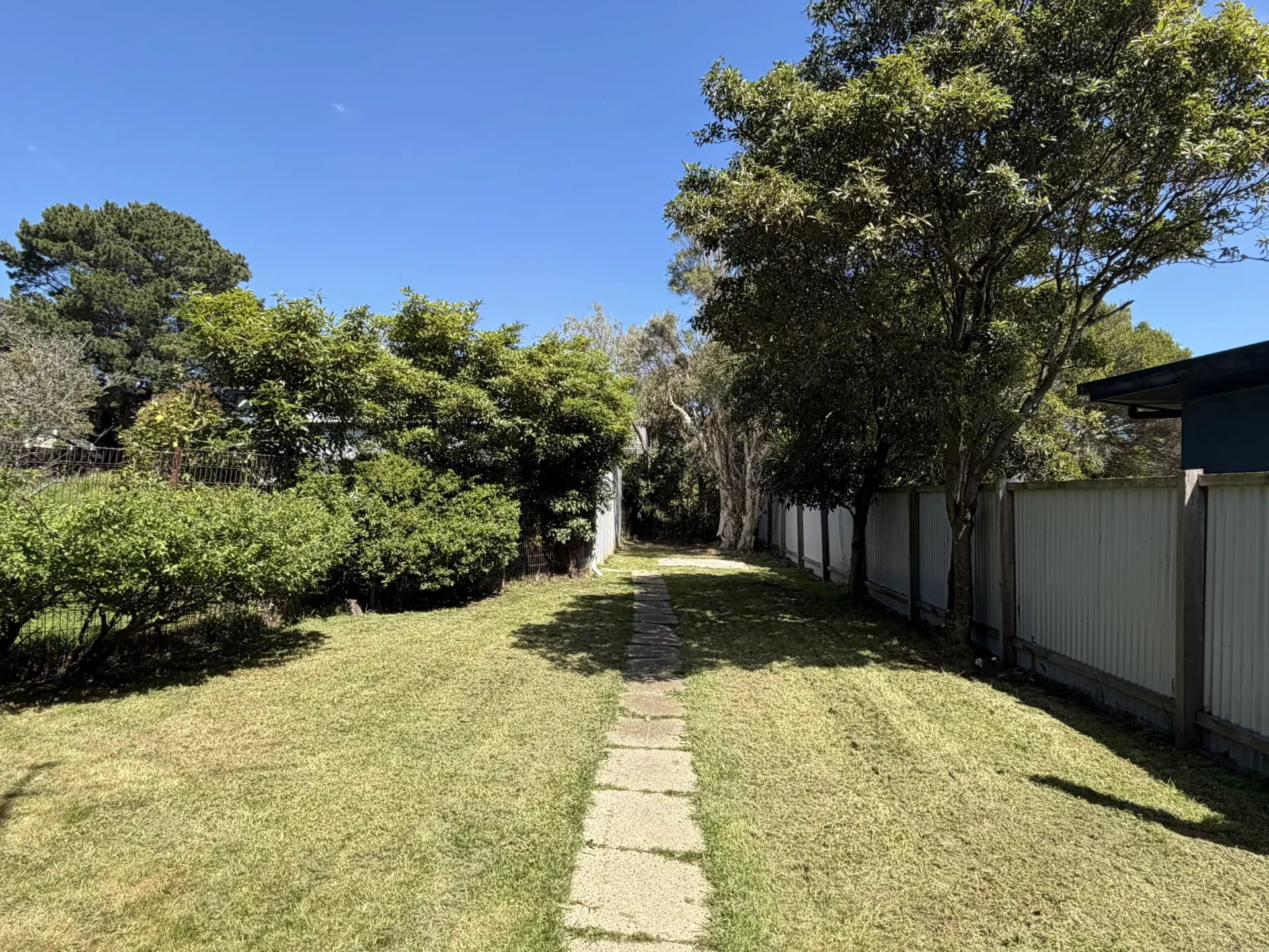 Long grassy rear yard with a stepping-stone path, trees and shrubs, bordered by fencing