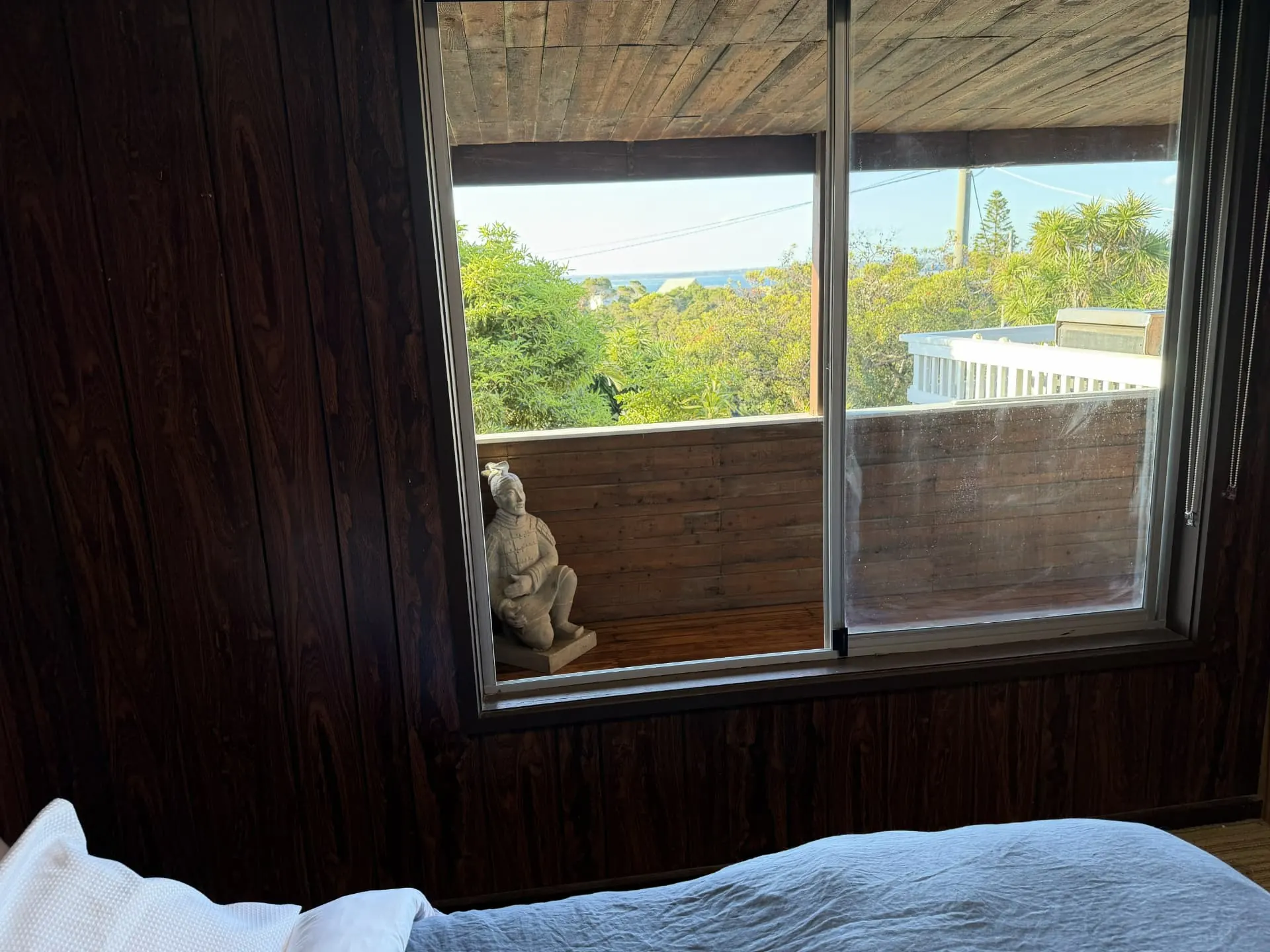 View from the bed in the upstairs double bedroom through a window to the covered balcony, a statue and treetops with water in the distance