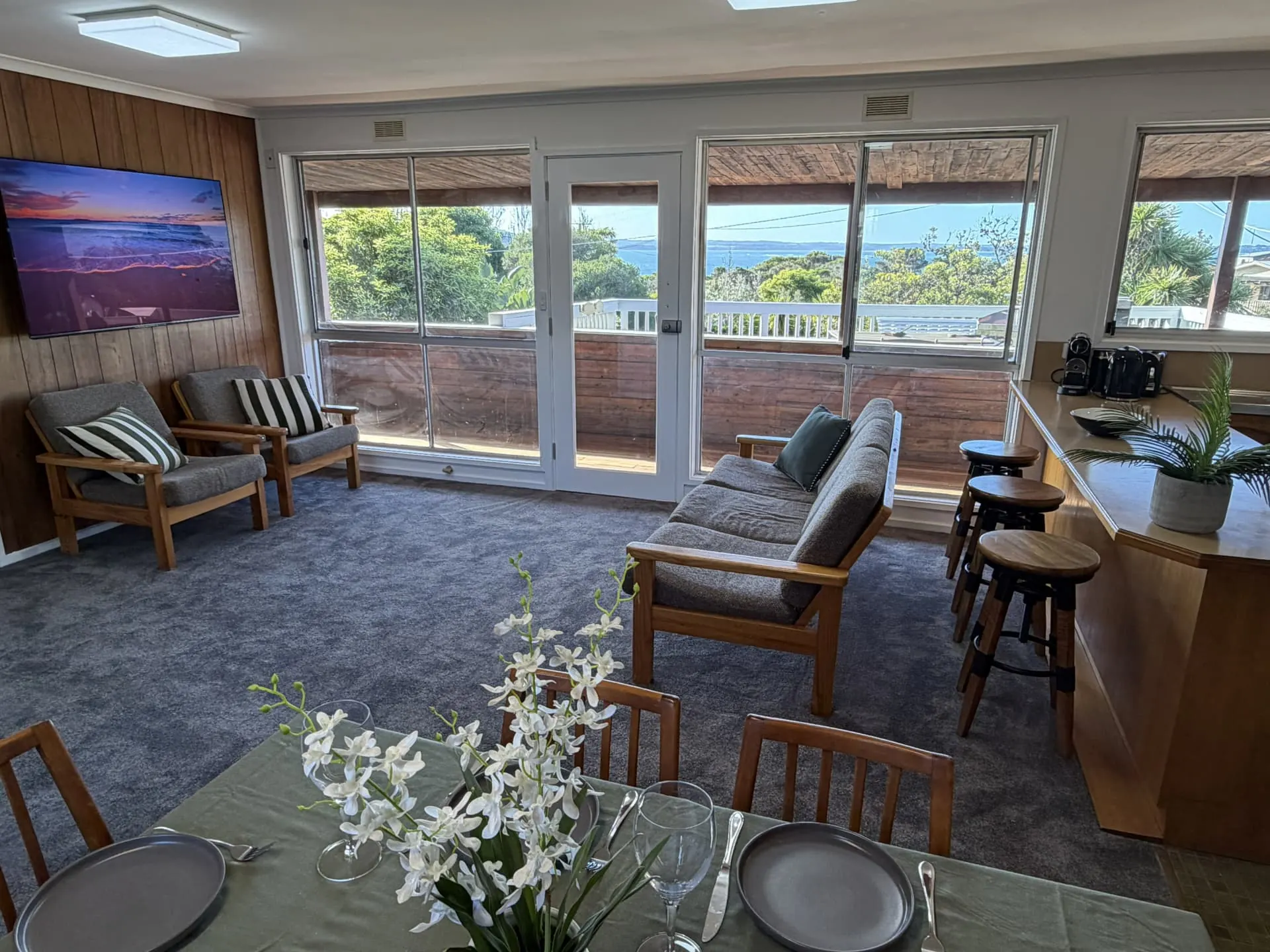 Upstairs lounge with timber armchairs, a sofa and a breakfast bar, with large windows and a glazed door opening to a covered balcony and water views beyond the trees
