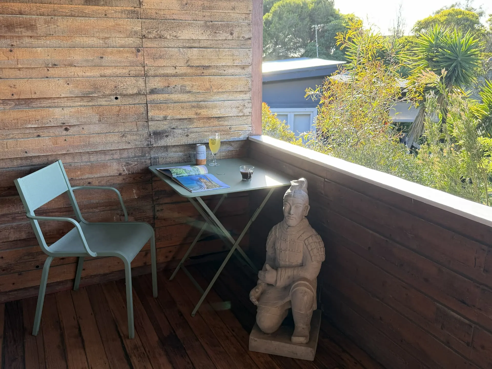 Covered timber balcony with a small table and chair, drinks and an open magazine, a statue and garden greenery beyond the balustrade