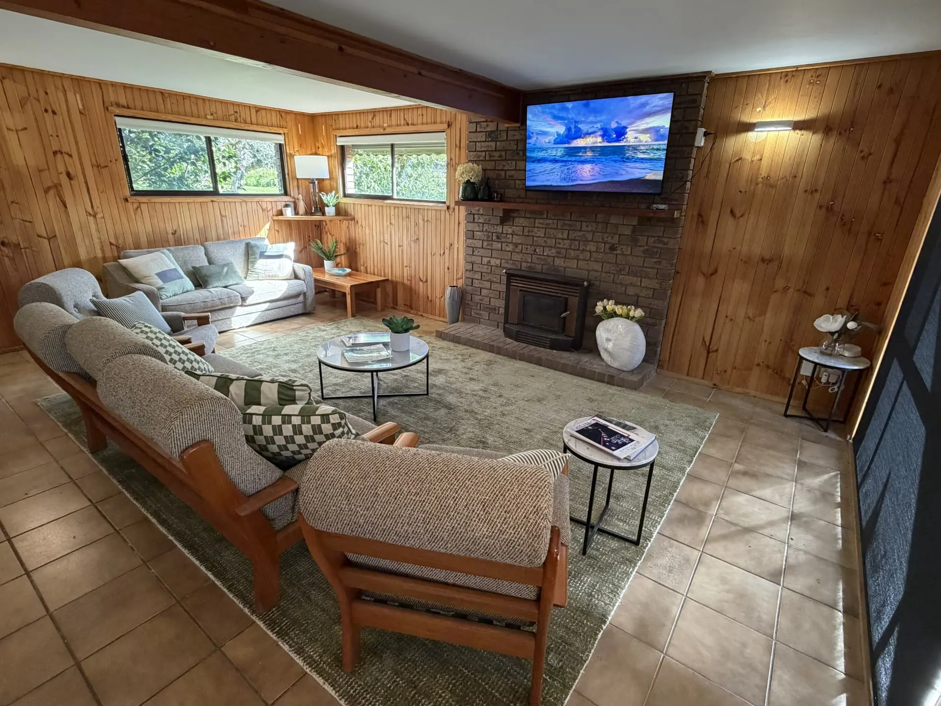 Downstairs living room with timber-panelled walls, sofas around a green rug, a brick fireplace with a heater and a wall-mounted TV