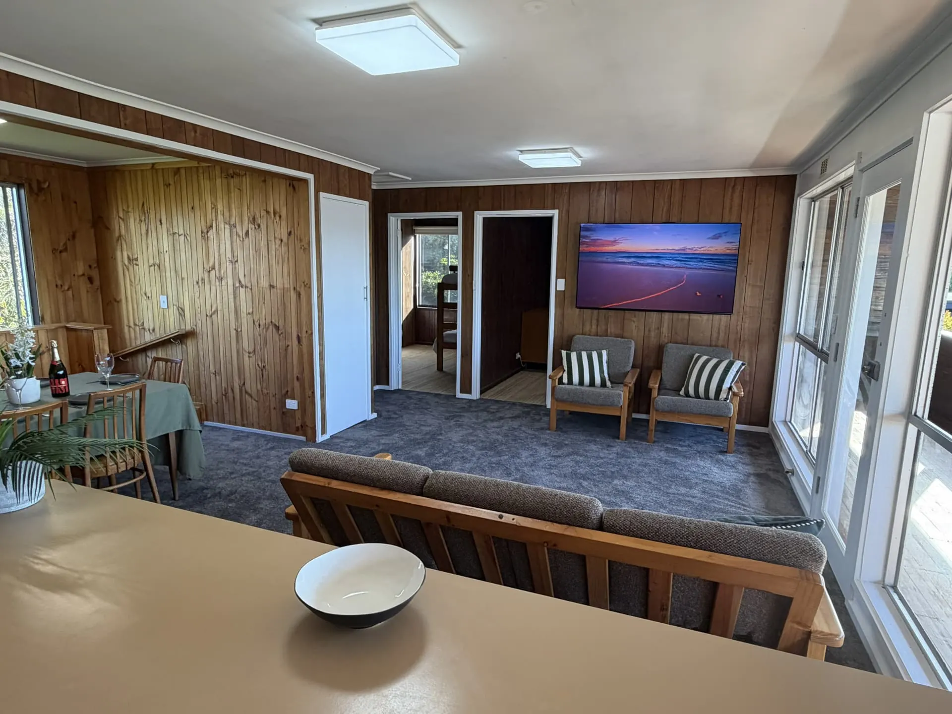 Open-plan upstairs lounge and dining area with a set dining table, timber-panelled walls, armchairs and a wall-mounted TV, seen from the kitchen bench
